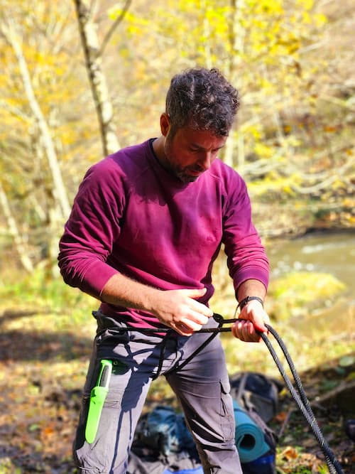 Man oefent touwtechnieken tijdens een survival- en bushcraftcursus in het bos, met zichtbaar mes en uitrusting, omringd door herfstbladeren bij een rivier.