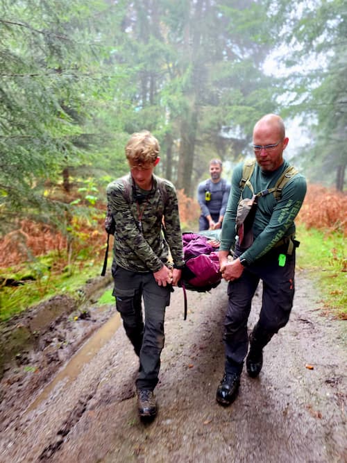 Drie mannen dragen een brancard over een modderig bospad tijdens een survivaloefening.