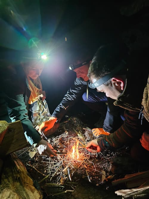Drie mensen maken vuur in het donker met hoofdlampen en droge takjes.