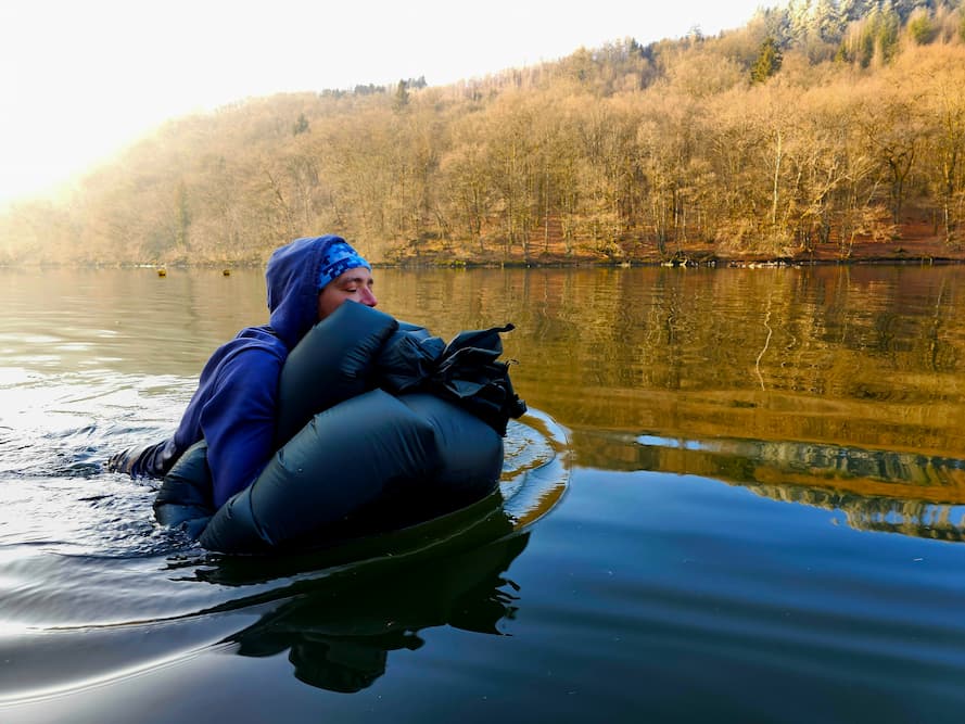 Een survivalist steekt een koud meer over door te zwemmen met uitrusting als drijfmiddel.