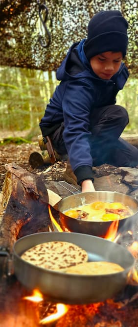 Een jonge jongen bakt eieren en platbrood boven een open vuur tijdens een bushcraftactiviteit.